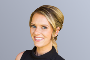 "Smiling woman with blonde hair in an updo, wearing a black sleeveless top, against a gray background."