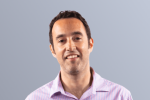 Man in a pink checkered shirt smiling against a light gray background.