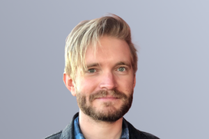 Man with blonde hair and beard in a denim jacket standing against a light gray background.