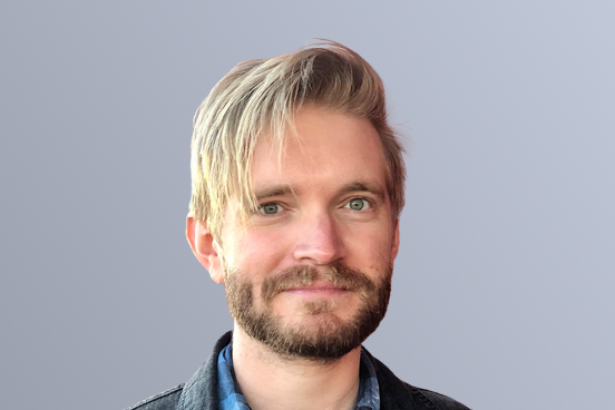 Man with blonde hair and beard in a denim jacket standing against a light gray background.