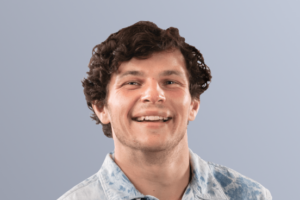 Man smiling at the camera with curly hair wearing a light blue patterned shirt against a light gray background.
