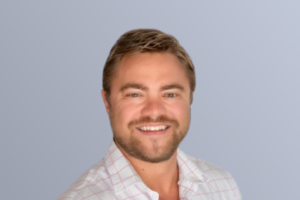 "Smiling man with short hair and beard wearing a light plaid shirt against a plain background."