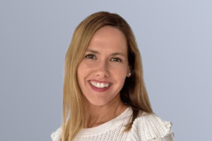 "Smiling person with light brown hair wearing a white knit sweater against a light gray background."