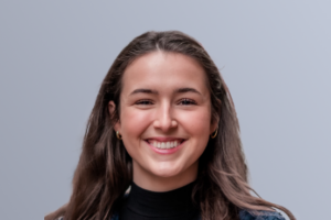 Smiling young woman with long brown hair, wearing a black top and jean jacket, against a light grey background.