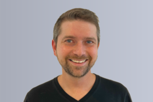 "Smiling man with brown hair wearing a patterned shirt against a plain background"