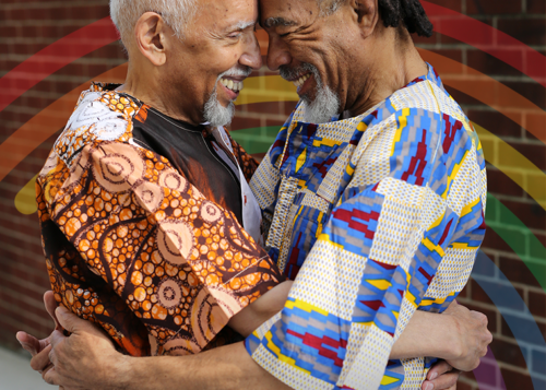 Two elderly men in vibrant, colorful traditional clothing embrace and smile warmly against a brick wall background.