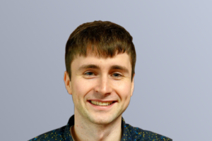 "Young man smiling against a plain light blue background, wearing a dark patterned shirt, short brown hair."