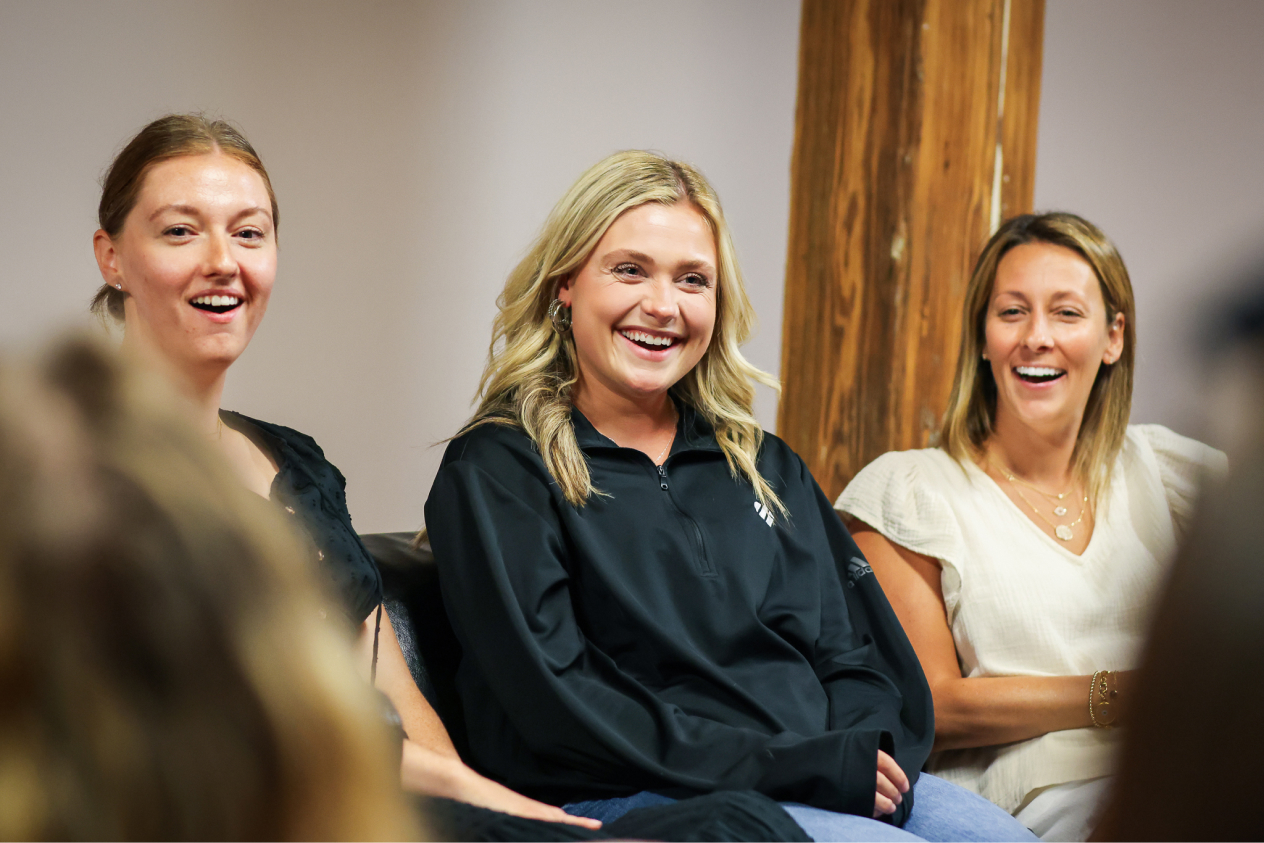 Three women sitting together and smiling in a casual indoor setting with a wooden pillar in the background