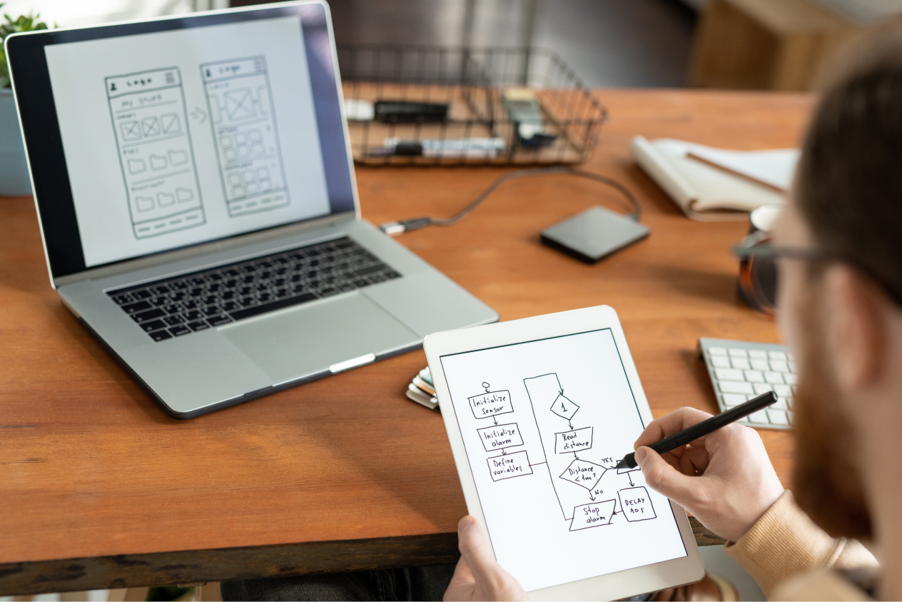 Person sketching wireframe design on a tablet with laptop displaying similar wireframe on the desk.