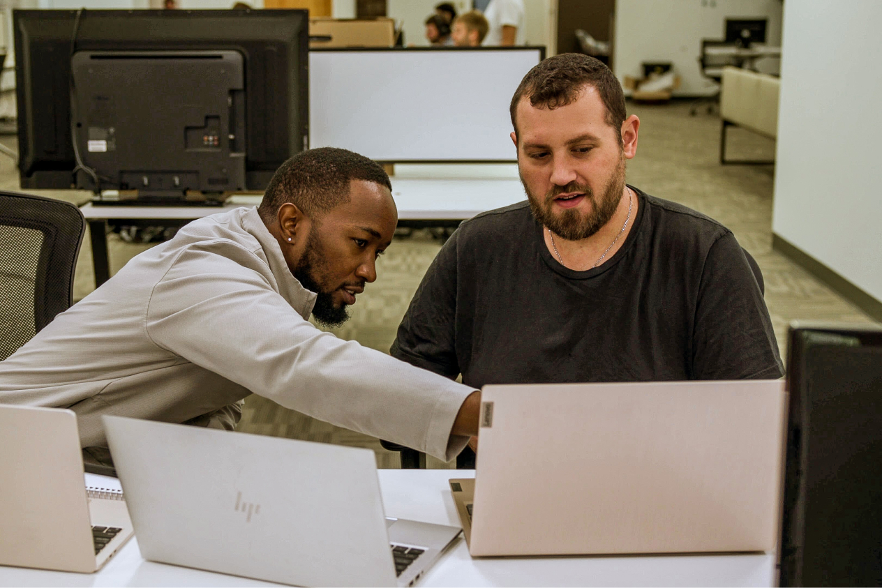 Two men collaborating at a table with laptops in an office environment, one man pointing at the screen.