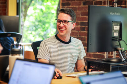 Man smiling and working on computer at desk with brick wall in background and greenery outside window.