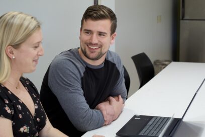 Two people sitting at a table with a laptop, engaged in conversation while smiling.