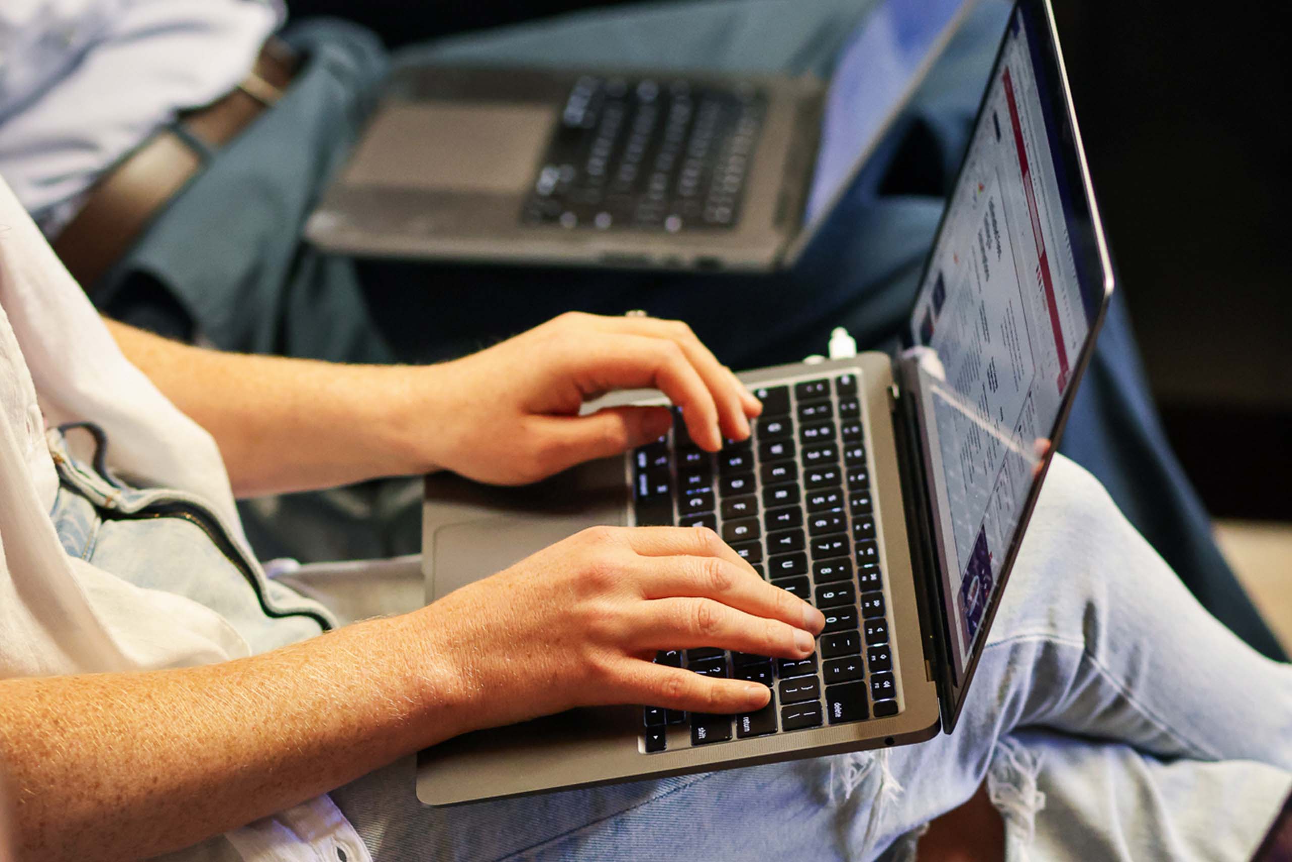 Person typing on a laptop while sitting, with another laptop visible in the background.