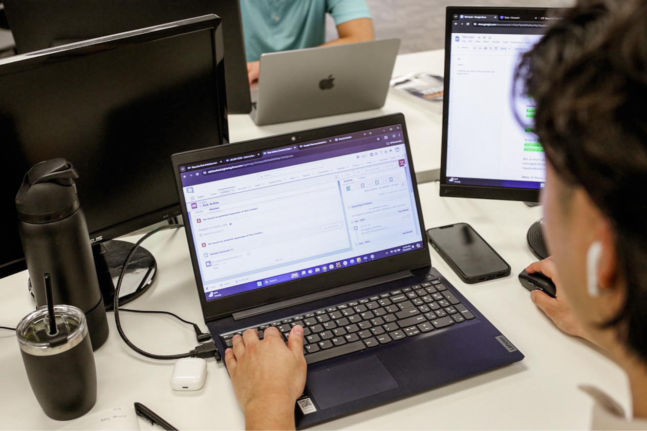 Person working on a laptop at a desk with multiple monitors and a smartphone, collaborating online with a team.