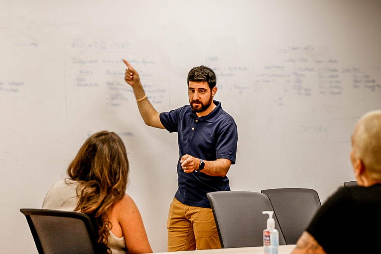 Man in a blue shirt giving a presentation to three people in a conference room, with a whiteboard in the background.