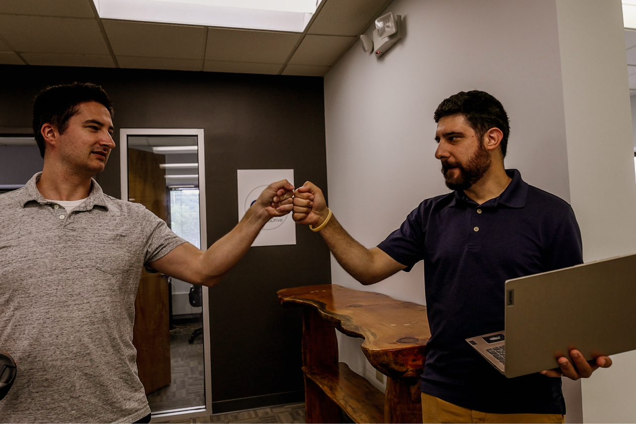 Two men fist bumping in an office hallway, one holding a laptop