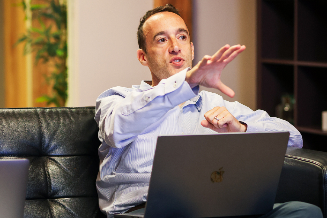 Man explaining something while sitting on a black leather couch, holding a laptop with an apple logo on his lap.