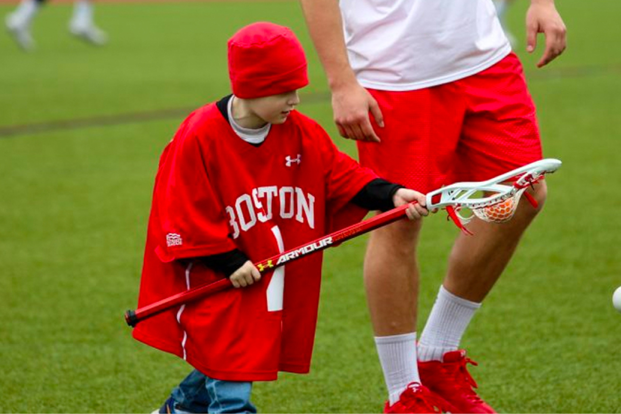 Young child playing lacrosse on a field wearing a red Boston jersey and matching hat