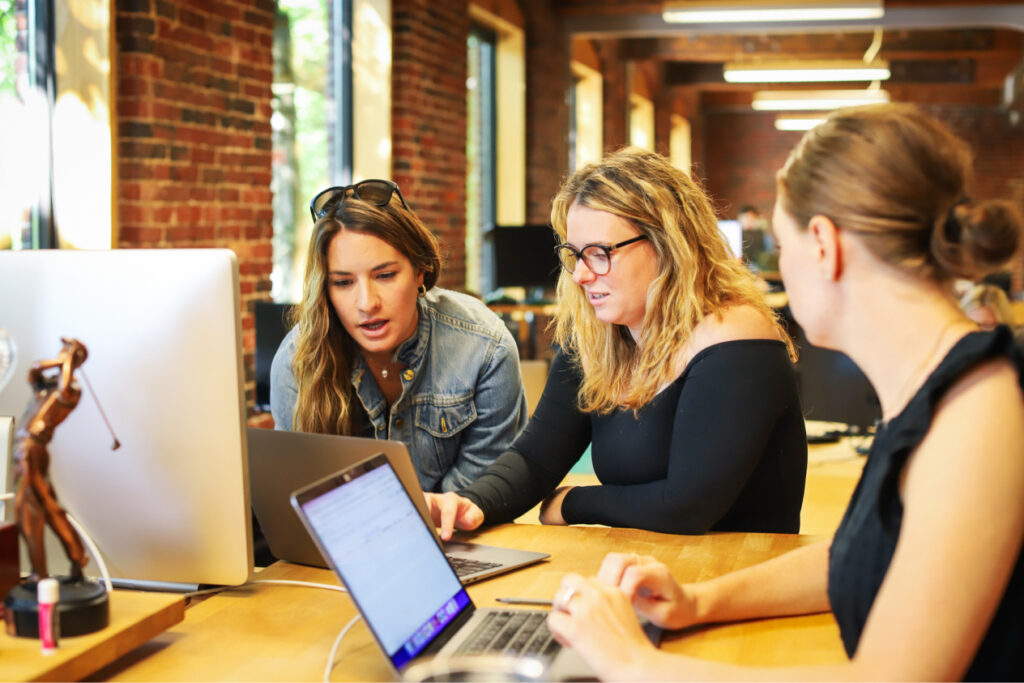 Women collaborating on a project in a modern office with laptops and a brick wall in the background