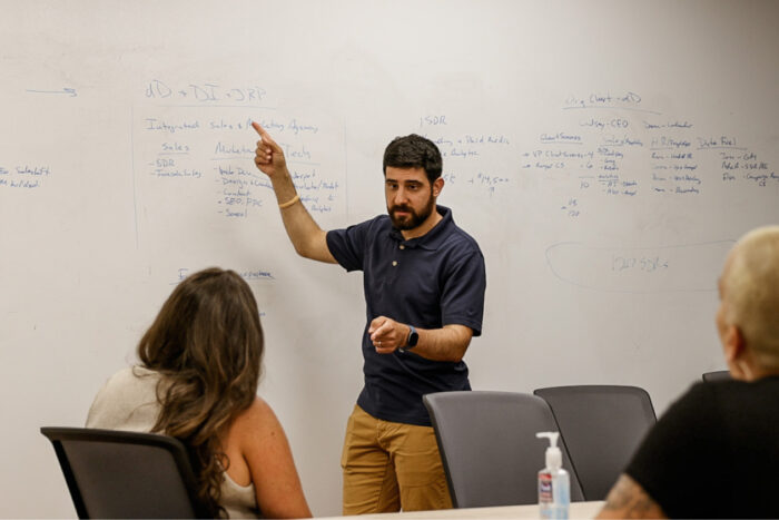 Person giving a presentation, pointing at notes on a whiteboard, while two people listen in a meeting room