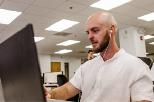 Bald man wearing a white shirt and earphones working on a laptop in a bright office environment.
