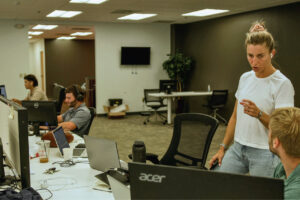Colleagues working in an office, some using computers while one person stands and talks to another who is sitting.