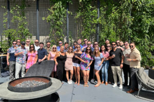 Group of people standing together outdoors on a sunny day next to a fire pit with greenery in the background