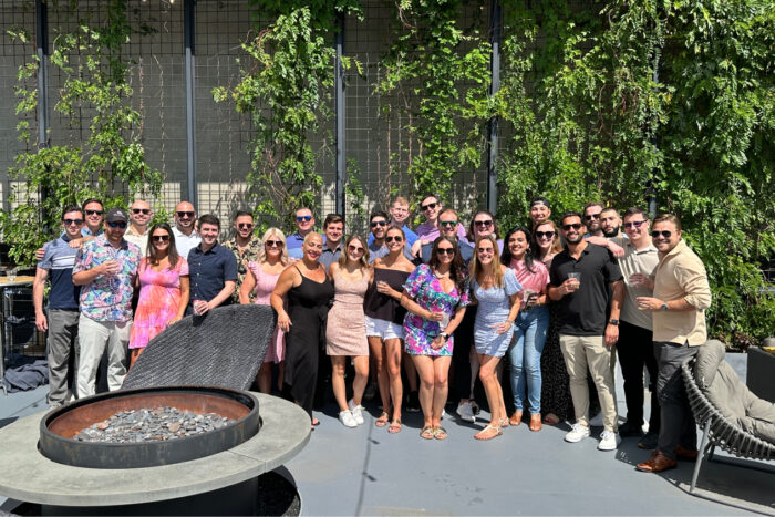 Group of people standing together outdoors on a sunny day next to a fire pit with greenery in the background