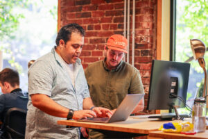 Two men collaborating on a project while working on a laptop at a modern office with exposed brick walls and sunlight.