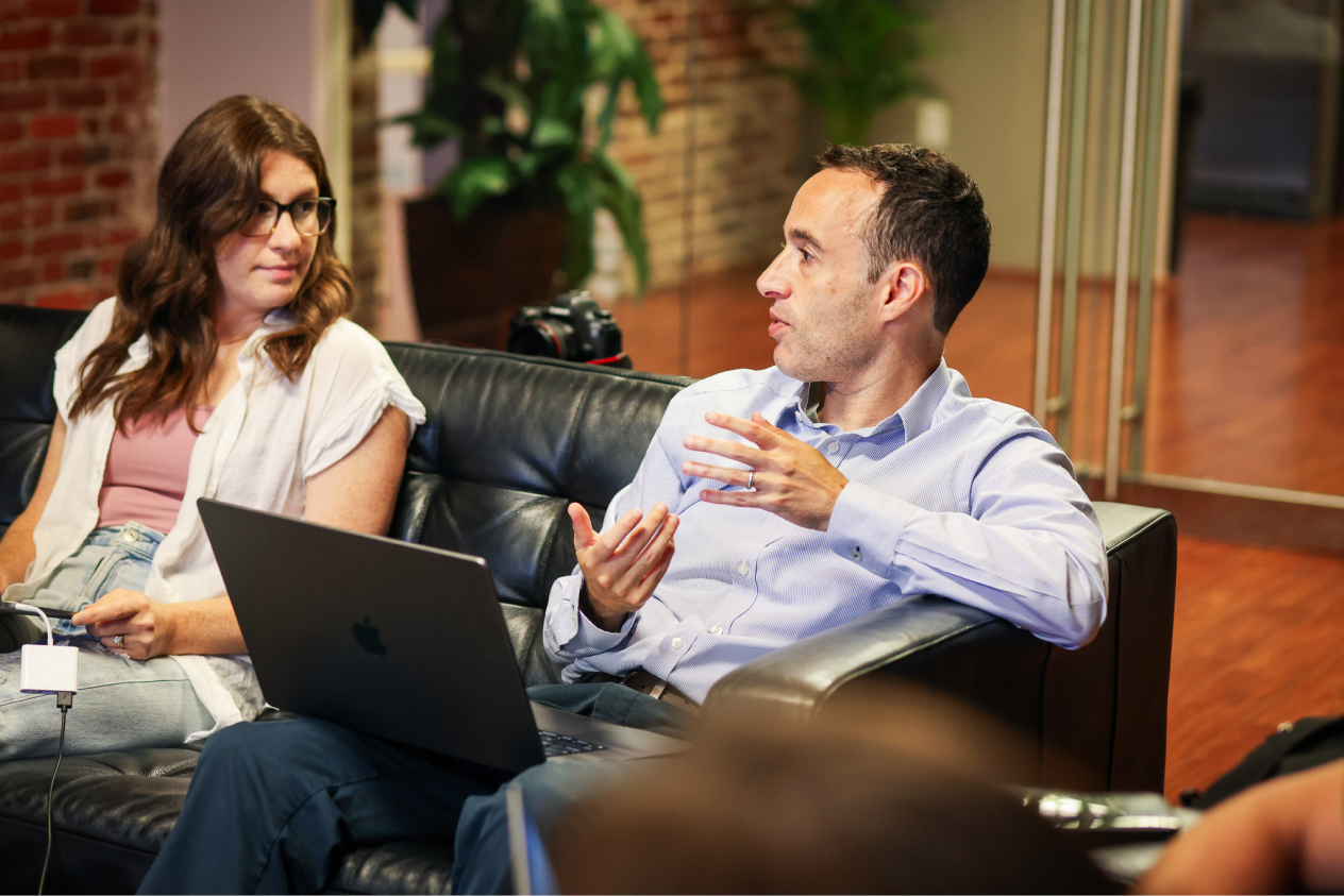Two colleagues discussing work while sitting on a black leather couch with a laptop in a modern office setting.