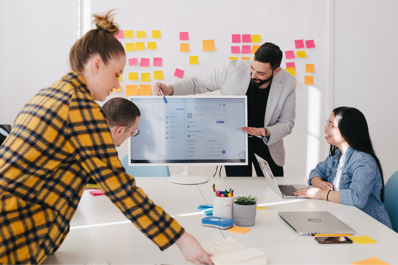 Team collaborating in a modern office with a large monitor, sticky notes on the wall, and laptops on the table