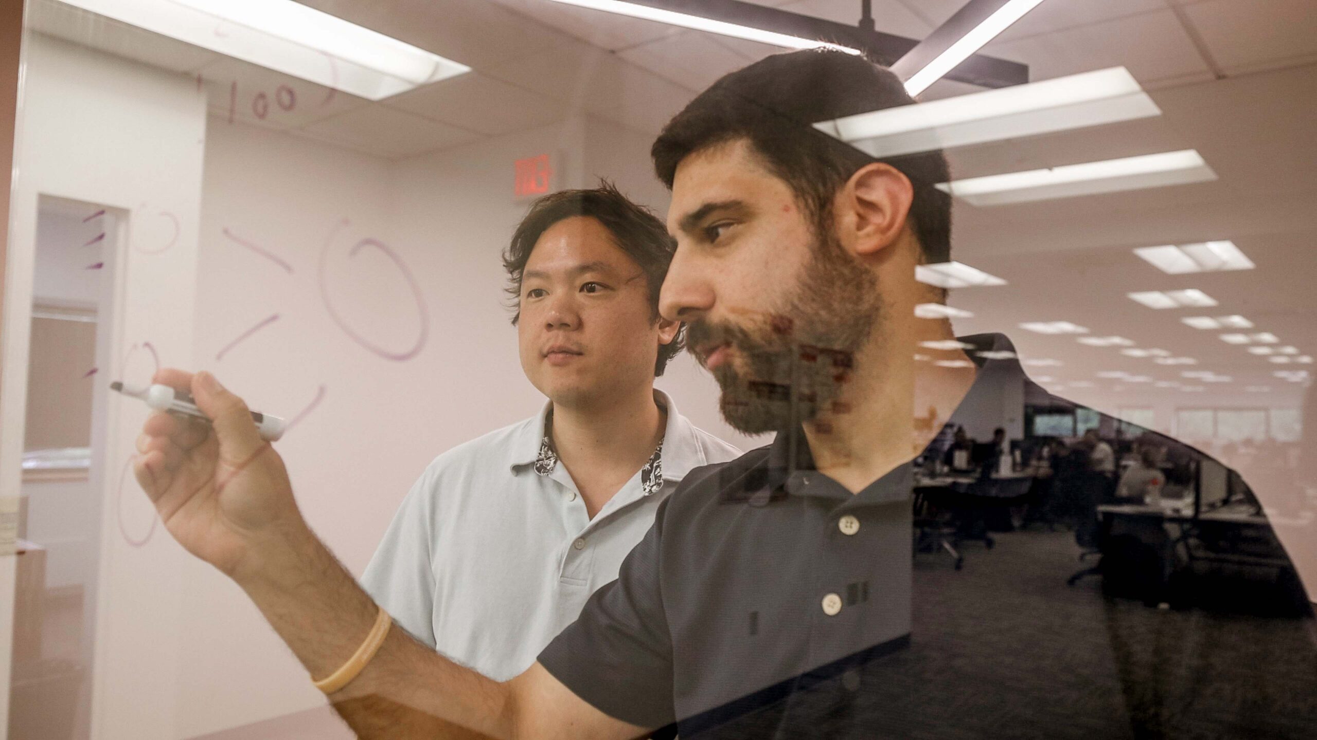 Two men collaborating and writing on a glass board in an office setting.