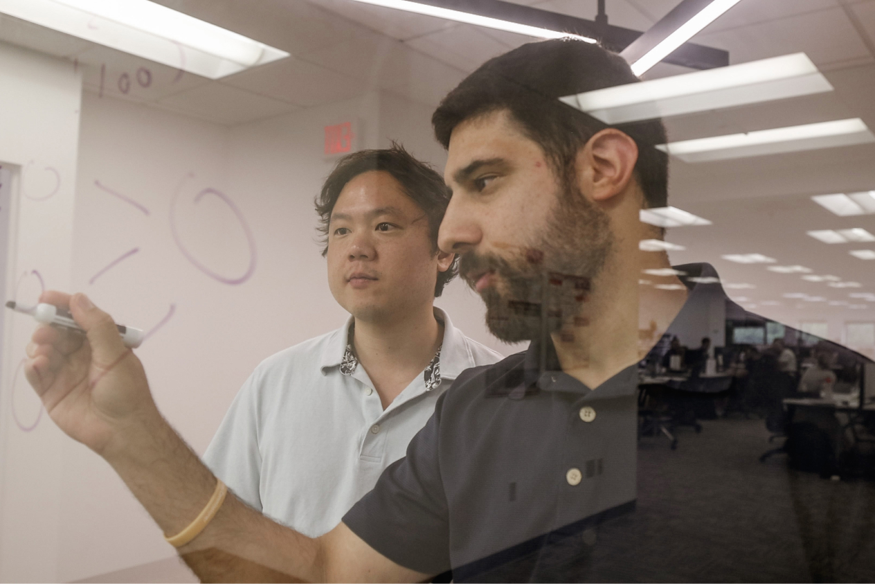 Two men brainstorming and writing on a glass board in a modern office setting.