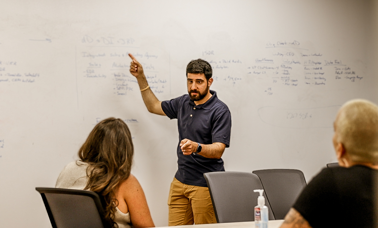 Man in a blue shirt pointing at a whiteboard during a meeting with three colleagues in an office setting