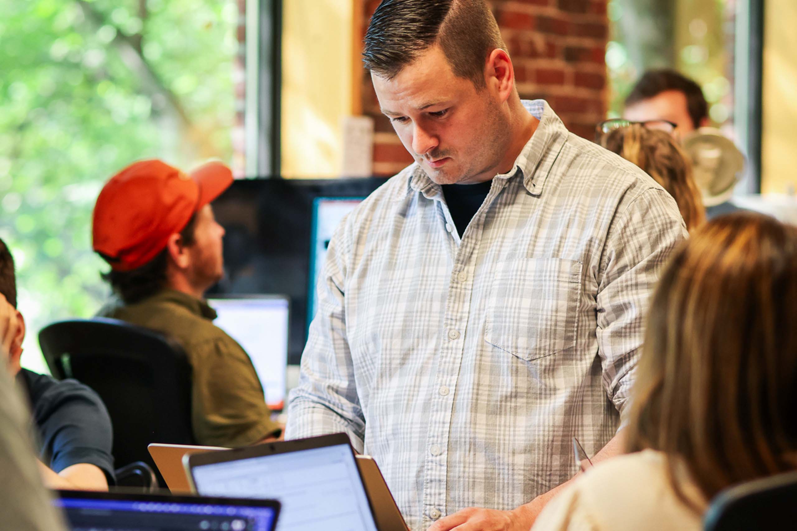 Man in a checked shirt working on a laptop in a busy office environment with colleagues in the background.