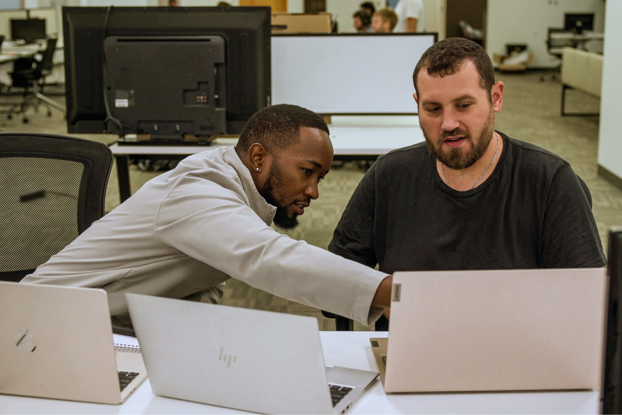 Two men in an office collaborating on laptops, focusing on a shared task.