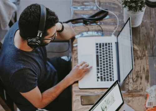 Man wearing headphones working on laptop at a wooden desk with another laptop and plant accessory nearby.