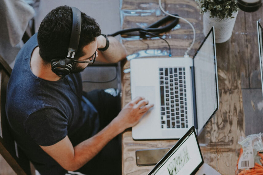 Man wearing headphones working on laptop at a wooden desk with another laptop and plant accessory nearby.