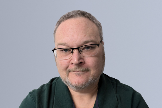 Man wearing glasses and a green shirt with a neutral expression against a gray background.