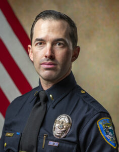 A police officer in uniform stands confidently in front of the American flag background.